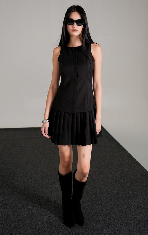 Rosemary Dress Black, gaun mini tanpa lengan warna hitam dengan kancing depan dan rok lipit pendek, dipadukan dengan kacamata hitam dan sepatu boots tinggi.
