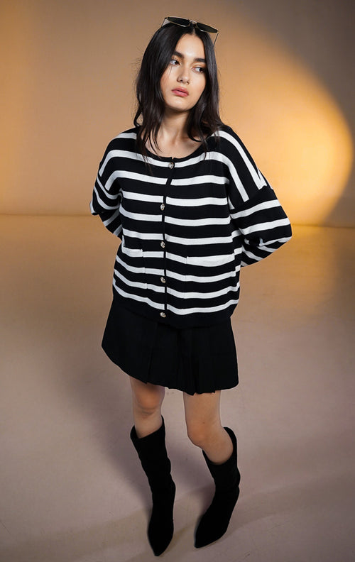 Model wanita memakai Saint Outer Black, cardigan rajut motif garis hitam putih dengan saku depan dan kancing emas, dipadukan dengan rok hitam pendek dan sepatu boots tinggi