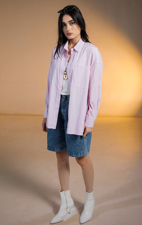 Model wanita memakai Sakuya Top Pink sebagai outer, kemeja motif garis vertikal pink putih lengan panjang dengan saku depan, dipadukan dengan celana pendek denim.