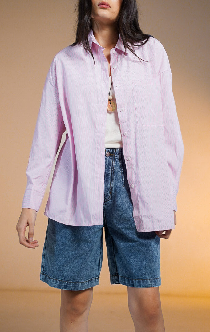 Model wanita memakai Sakuya Top Pink sebagai outer, kemeja motif garis vertikal pink putih lengan panjang dengan saku depan, dipadukan dengan celana pendek denim.