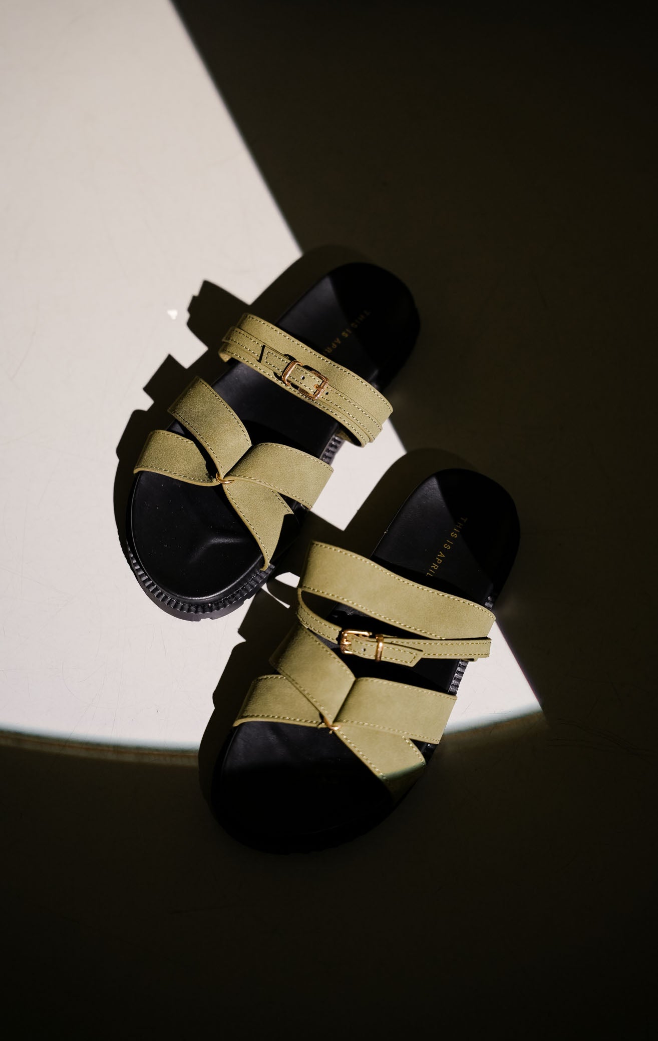 Tampilan flat lay sepasang Violet Sandals warna olive, sandal flat teplek wanita tali menyilang bahan suede hijau zaitun dengan gesper emas dan sol tebal gerigi hitam dari This Is April.