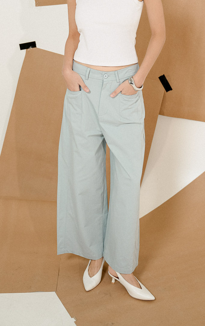 Model berdiri dengan tangan di saku mengenakan Sapphire Straight Leg Pants Light Blue, produk celana panjang wanita potongan lurus warna biru muda dengan detail kancing depan. 