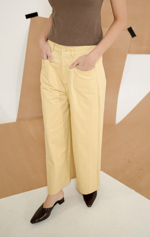 Model berdiri dengan tangan di saku mengenakan Sapphire Straight Leg Pants Yellow, produk celana panjang wanita potongan lurus warna kuning lembut dengan detail kancing depan. 