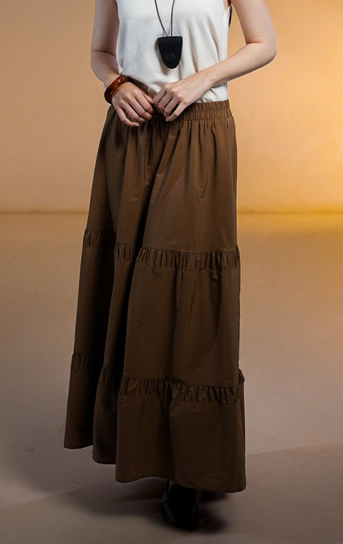 Model wanita memakai Seamus Skirt Brown, rok maxi panjang warna coklat tua dengan detail sambungan rempel bertingkat (tiered) dipadukan dengan atasan putih.