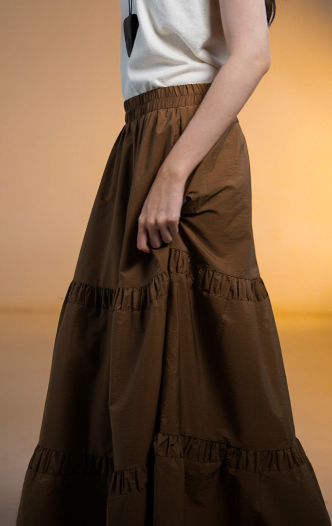 Model wanita memakai Seamus Skirt Brown, rok maxi panjang warna coklat tua dengan detail sambungan rempel bertingkat (tiered) dipadukan dengan atasan putih.