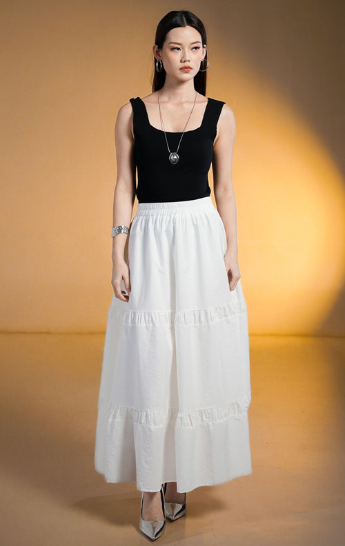 Model wanita memakai Seamus Skirt White, rok maxi panjang warna putih dengan detail sambungan rempel bertingkat (tiered) dan pinggang karet, dipadukan dengan tank top hitam.