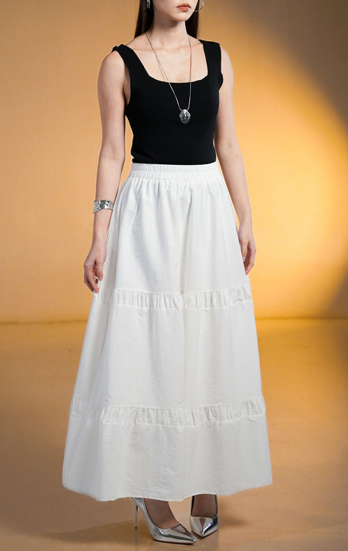 Model wanita memakai Seamus Skirt White, rok maxi panjang warna putih dengan detail sambungan rempel bertingkat (tiered) dan pinggang karet, dipadukan dengan tank top hitam.