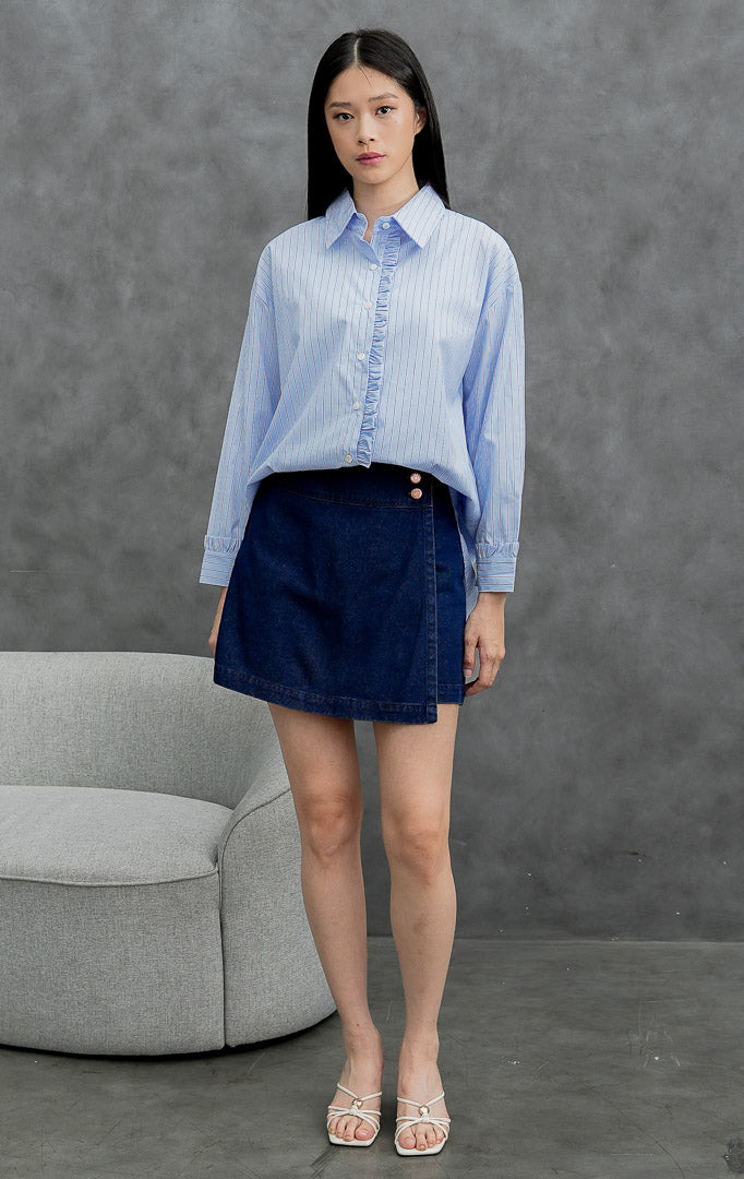 Kemeja katun wanita lengan panjang warna biru muda (light blue) Sevran Top berkerah dengan motif garis vertikal (vertical stripes), detail rumbai (ruffle trim), dan bukaan kancing depan.