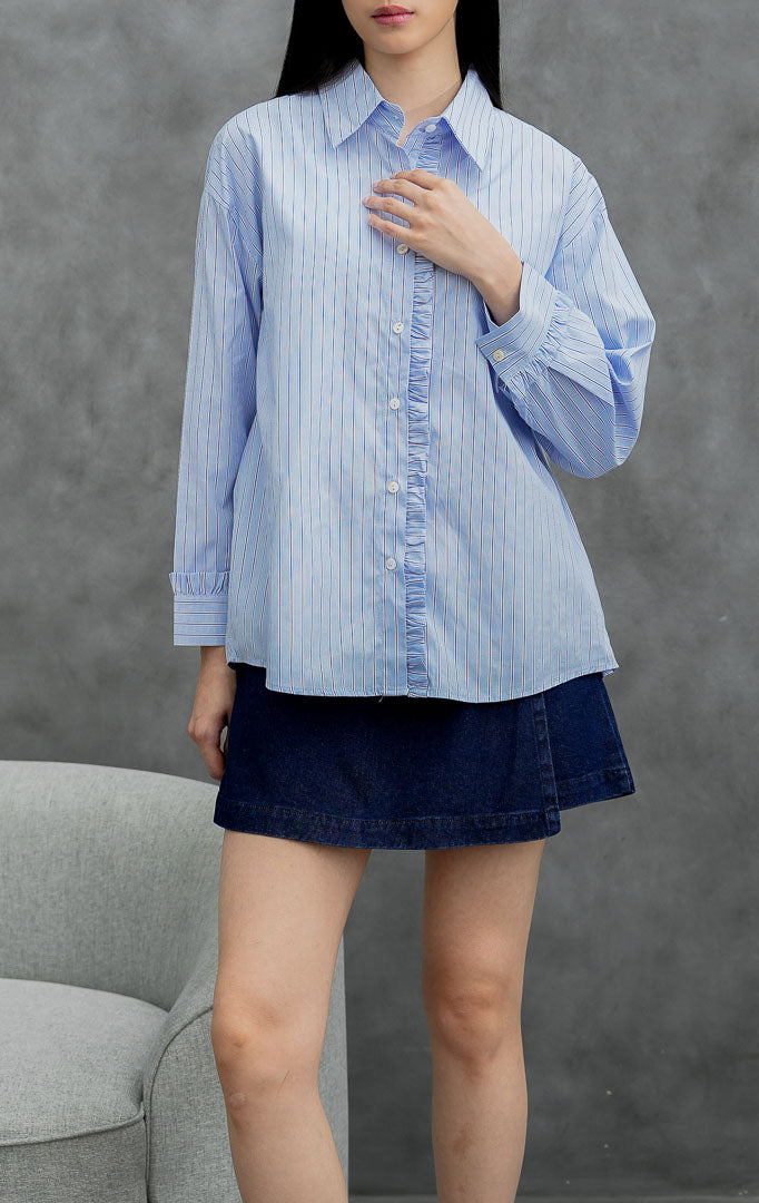 Kemeja katun wanita lengan panjang warna biru muda (light blue) Sevran Top berkerah dengan motif garis vertikal (vertical stripes), detail rumbai (ruffle trim), dan bukaan kancing depan.