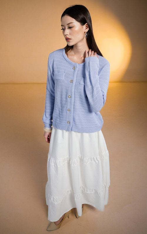 Model wanita memakai Shiloh Outer Blue, cardigan rajut lengan panjang warna biru periwinkle dengan kancing emas dan saku, dipadukan dengan rok putih
