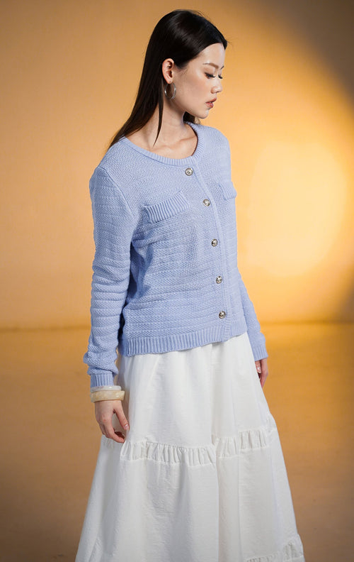 Model wanita memakai Shiloh Outer Blue, cardigan rajut lengan panjang warna biru periwinkle dengan kancing emas dan saku, dipadukan dengan rok putih