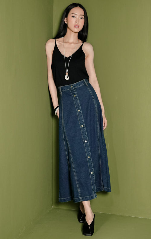 Rok panjang wanita Solde Skirt berbahan denim warna biru gelap (dark denim) dengan siluet A-line, dilengkapi detail jahitan panel vertikal yang rapi, tempat sabuk (belt loops), dan deretan kancing metal penuh di bagian tengah depan.