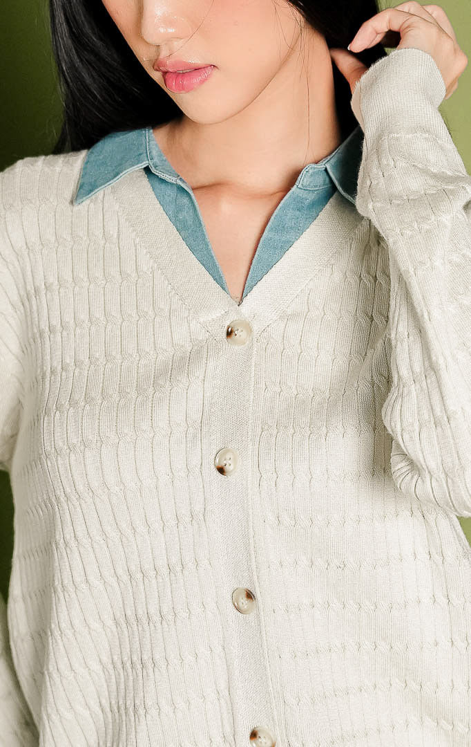 Kardigan rajut wanita Solene Outer lengan panjang warna ivory, memiliki tekstur rajut pola kepang vertikal, dilengkapi aksen lapisan kerah bahan denim biru kontras pada bagian leher V-neck, serta deretan kancing depan bernuansa cokelat.