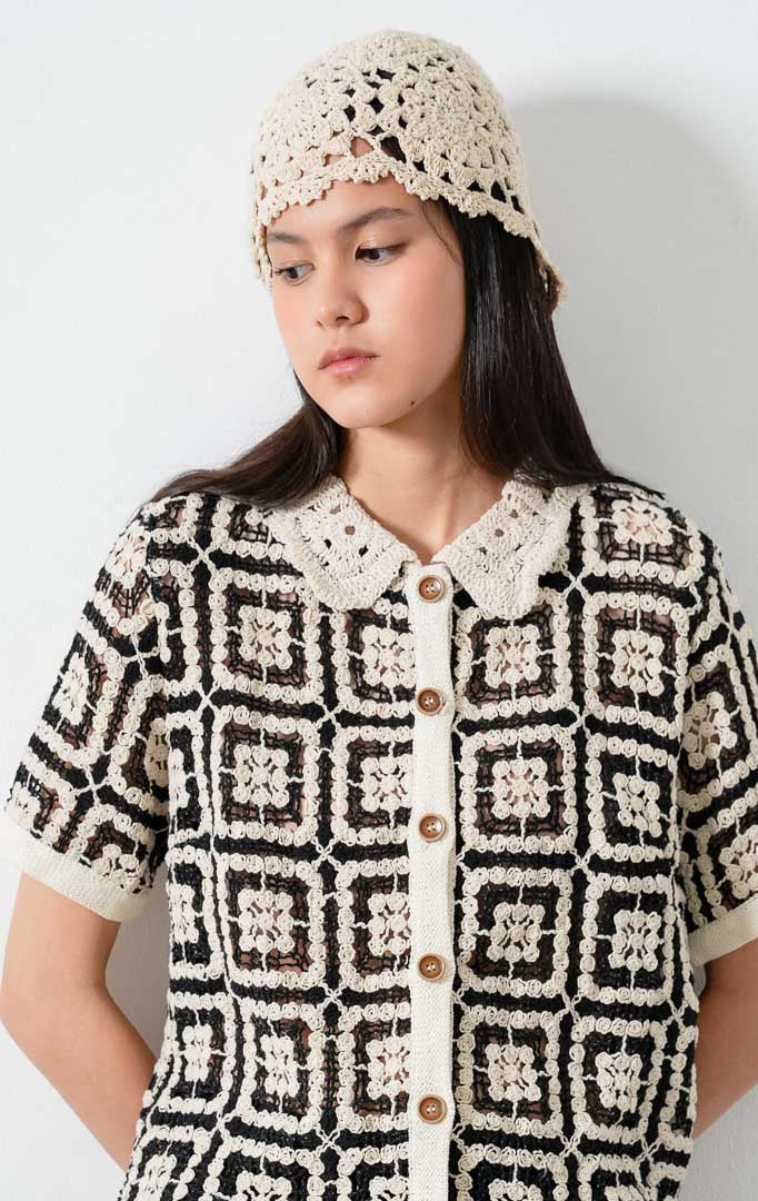 Atasan rajut motif geometris warna hitam (black crochet top) Sorae Top dengan kerah model Peter Pan aksen rajut (crochet-inspired Peter Pan collar), lengan pendek, siluet santai (relaxed silhouette), dan bukaan kancing depan (button-down front). 