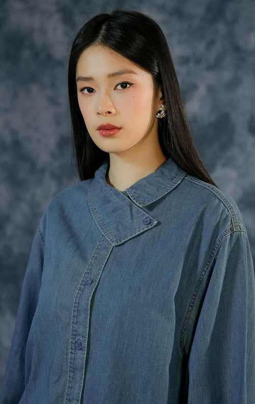 Kemeja lengan panjang wanita Steya berbahan denim dengan detail kerah asimetris yang unik (statement collar), kancing depan aktif, dan lengan kancing.
