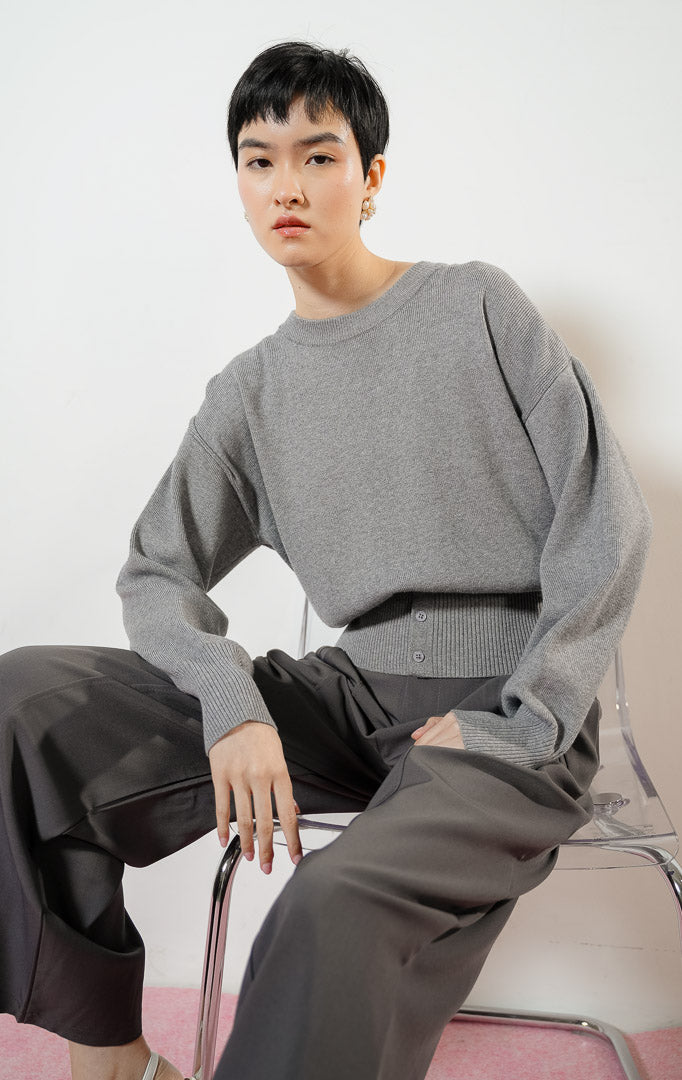 Model wanita mengenakan Cerise Button Hem Knit Sweater warna grey, atasan sweater rajut wanita lengan panjang berwarna abu-abu dengan karet pinggang rib lebar berdetail kancing. 