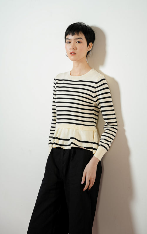 Model wanita mengenakan Season Striped Ruffle Hem Knit Sweater warna ivory, atasan sweater baju rajut wanita lengan panjang motif garis salur putih hitam dengan detail kelim peplum rumbai yang feminin. 