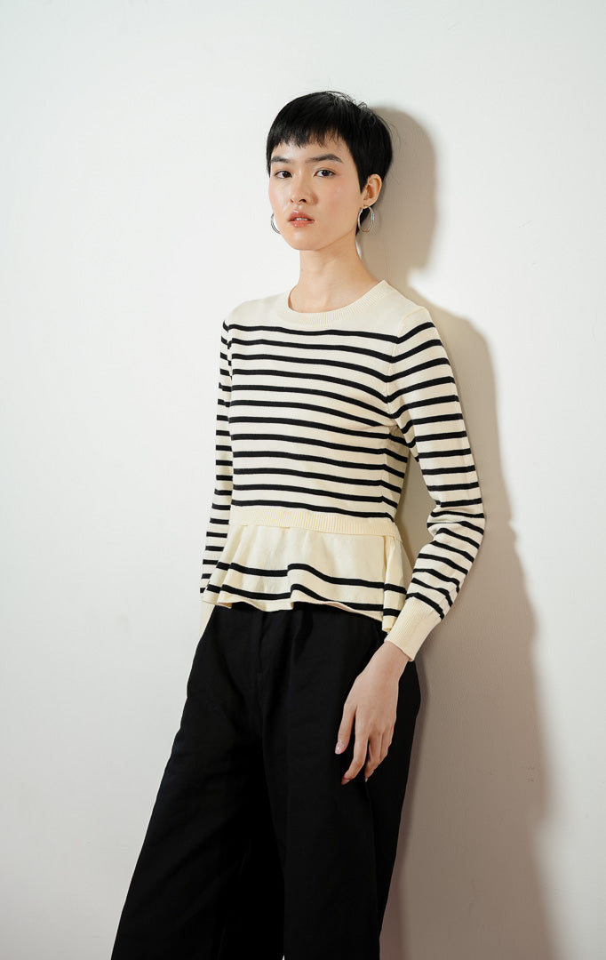 Model wanita mengenakan Season Striped Ruffle Hem Knit Sweater warna ivory, atasan sweater baju rajut wanita lengan panjang motif garis salur putih hitam dengan detail kelim peplum rumbai yang feminin. 