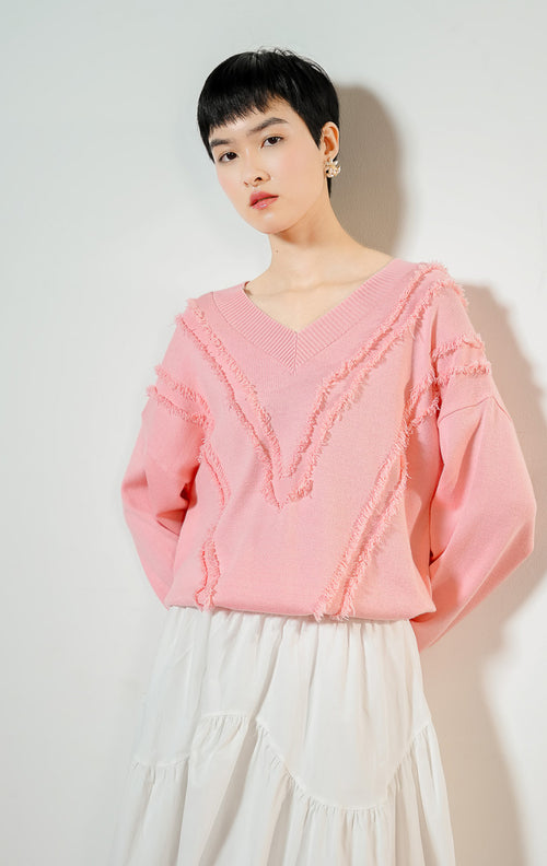 Model wanita mengenakan Wyetta V-Neck Fringe Knit Sweater warna pink, baju sweater rajut wanita leher V warna merah muda pastel dengan detail aksen jumbai rumbai bertekstur modis.