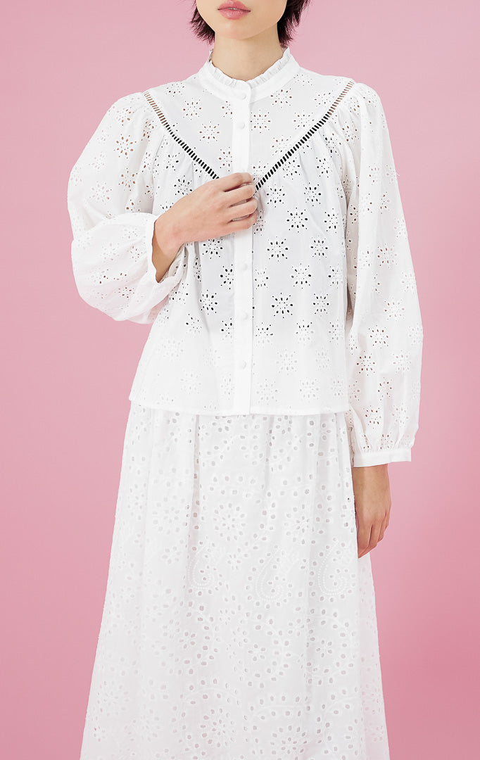 Atasan blus wanita Sylvia Top warna putih dari This Is April dengan detail bordir eyelet (lubang), kerah rempel (frill), dan kancing depan, dipadukan dengan rok bordir senada.