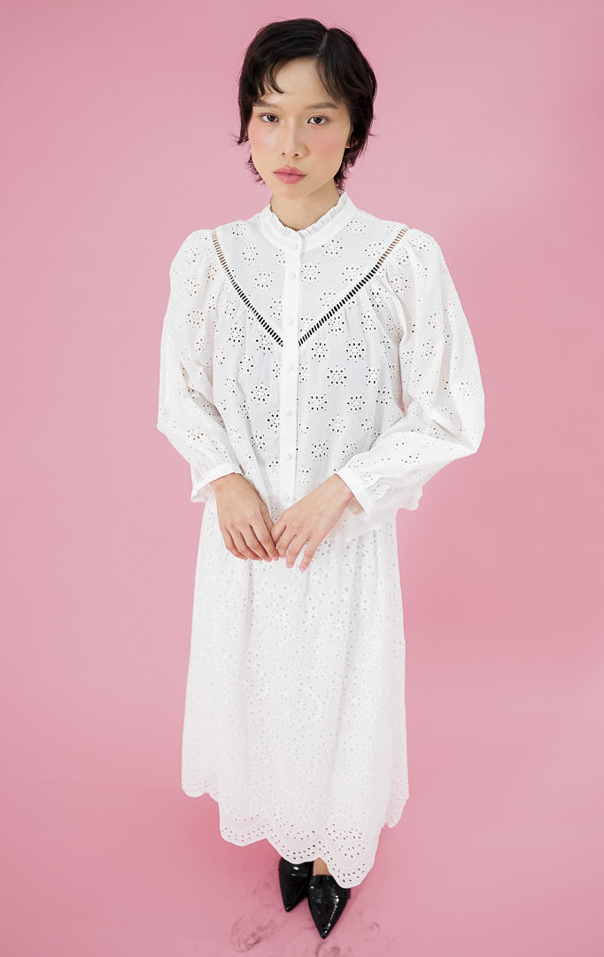 Atasan blus wanita Sylvia Top warna putih dari This Is April dengan detail bordir eyelet (lubang), kerah rempel (frill), dan kancing depan, dipadukan dengan rok bordir senada.