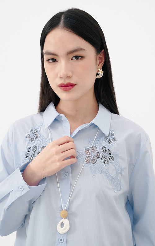 Close-up model mengenakan Tessa Top warna baby blue, kemeja katun lengan panjang dengan detail bordir bunga lace transparan yang cantik di sisi kanan dan kiri dada.
