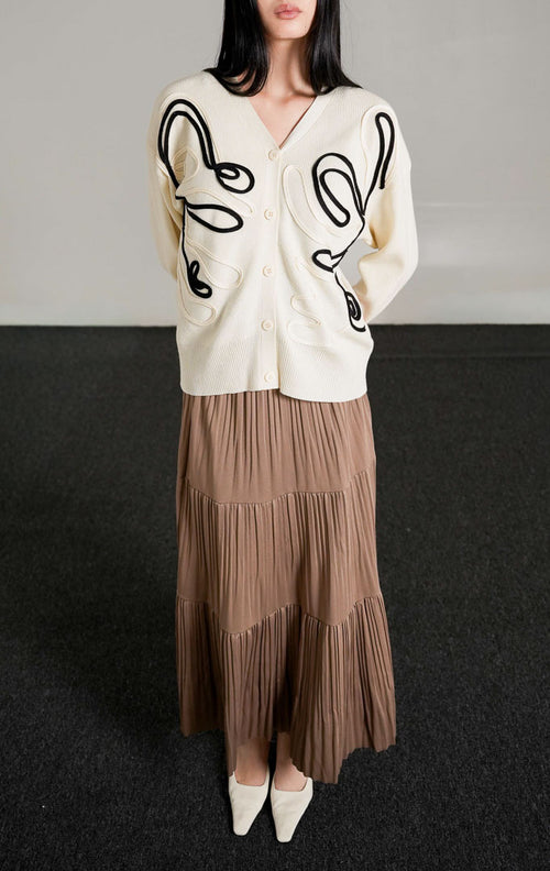Thea Patterned Knit Cardigan, kardigan rajut warna broken white dengan motif garis melengkung hitam, dipadukan dengan rok panjang cokelat.