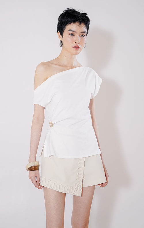 Tampak depan This Is April Airana Top warna putih, atasan wanita berbahan rajut dengan desain kerah drape berlipat dan detail aksen bros emas di bagian samping pinggang. 