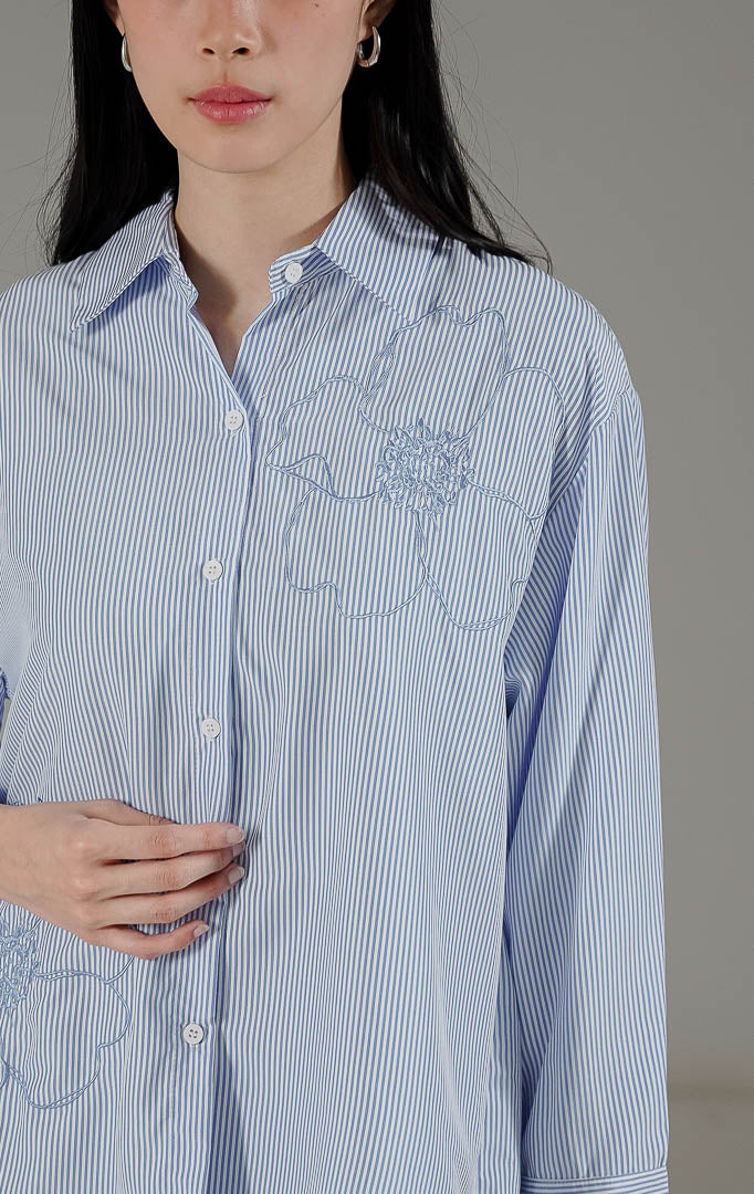 Amour Top Blue dari This Is April, kemeja lengan panjang motif garis warna biru klasik dengan detail bordir bunga besar dan potongan asimetris.