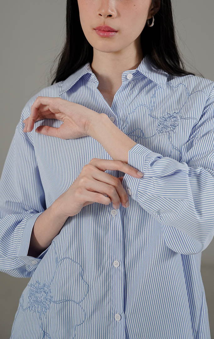 Amour Top Blue dari This Is April, kemeja lengan panjang motif garis warna biru klasik dengan detail bordir bunga besar dan potongan asimetris.