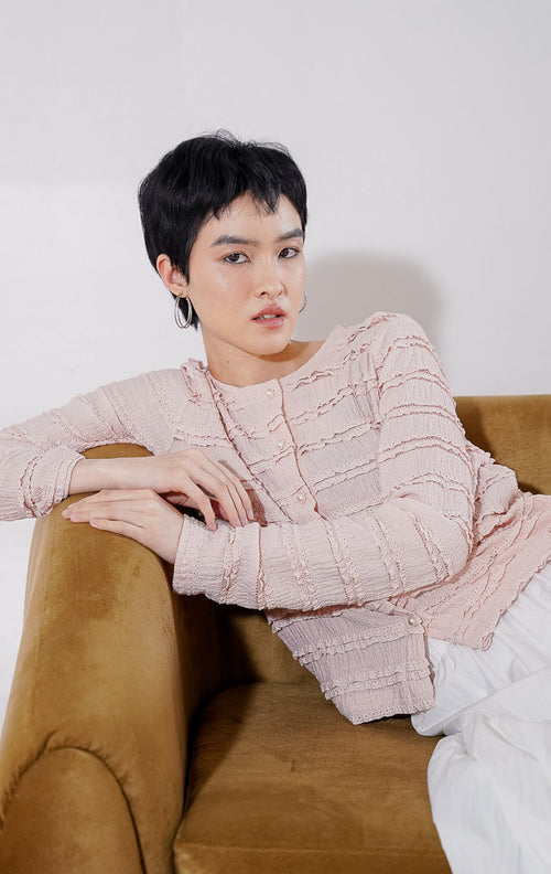 Outer crop wanita lengan panjang Anandra Top dari This Is April berwarna dusty pink dengan detail tekstur ruffle renda horizontal berlapis, kancing depan mutiara, dan material sheer menerawang, digantung di rak pakaian 