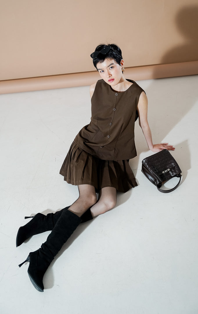 Cayden Dress Dark Brown dari This Is April, gaun tanpa lengan warna cokelat tua dengan detail rok lipit (pleated) bertumpuk dan kancing depan.