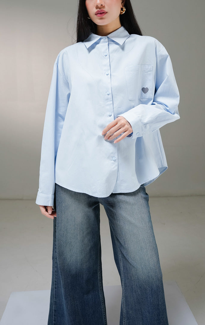 Kemeja basic lengan panjang wanita This Is April Claudette Top warna biru muda (light blue), dilengkapi satu saku di dada kiri dengan detail bordiran bentuk hati kecil warna biru tua. 