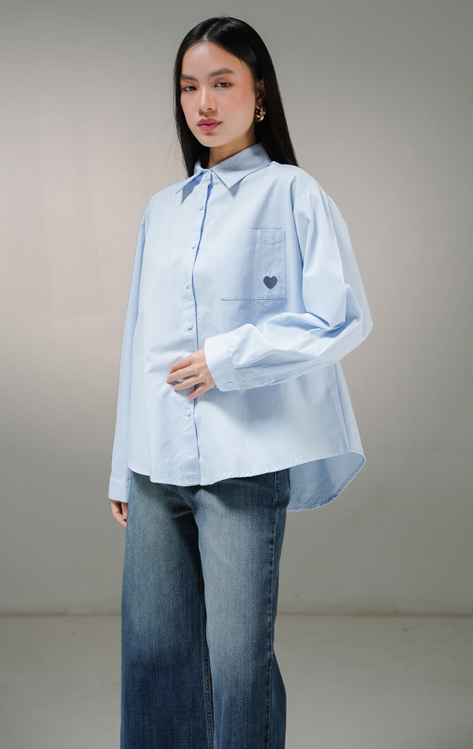 Kemeja basic lengan panjang wanita This Is April Claudette Top warna biru muda (light blue), dilengkapi satu saku di dada kiri dengan detail bordiran bentuk hati kecil warna biru tua. 