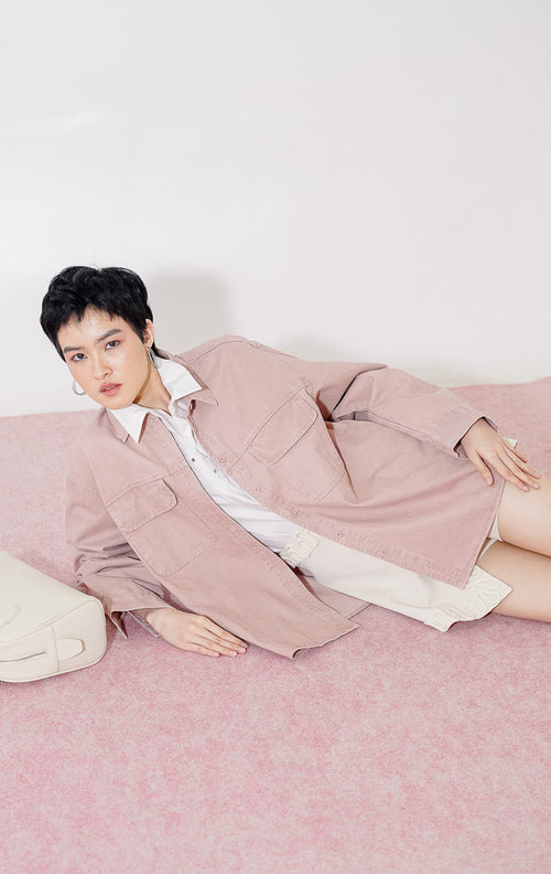 Kemeja wanita lengan panjang Dahlia Top dari This Is April berwarna dusty pink berbahan katun twill dengan dua kantong dada fungsional dan kancing depan, digantung di rak pakaian. 