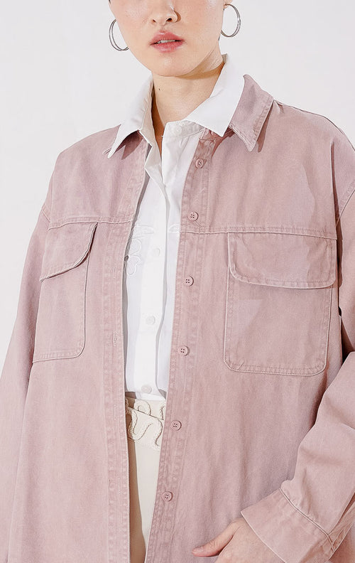 Kemeja wanita lengan panjang Dahlia Top dari This Is April berwarna dusty pink berbahan katun twill dengan dua kantong dada fungsional dan kancing depan, digantung di rak pakaian. 