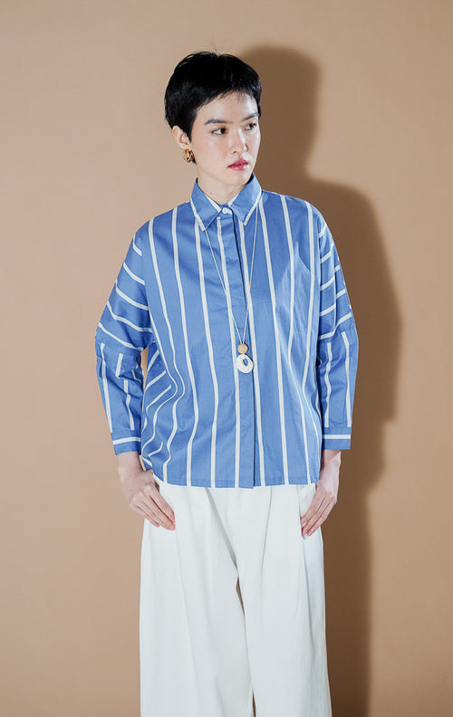 Deliora Top Blue dari This Is April, kemeja katun lengan panjang siluet boxy warna biru dengan motif garis vertikal putih dan kancing depan.
