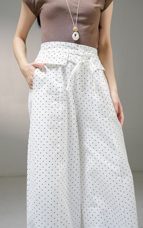 Celana panjang kulot wanita This Is April Evelyna Pants warna putih motif polkadot hitam kecil, dilengkapi kancing depan, saku penutup dekoratif, dan tali ikat pinggang. 