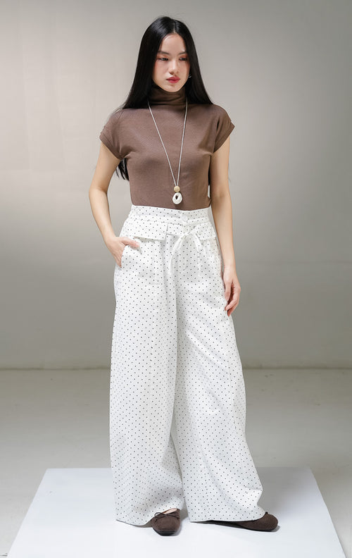 Celana panjang kulot wanita This Is April Evelyna Pants warna putih motif polkadot hitam kecil, dilengkapi kancing depan, saku penutup dekoratif, dan tali ikat pinggang. 