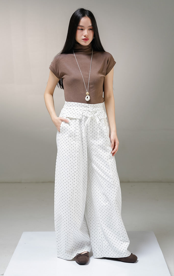 Celana panjang kulot wanita This Is April Evelyna Pants warna putih motif polkadot hitam kecil, dilengkapi kancing depan, saku penutup dekoratif, dan tali ikat pinggang. 
