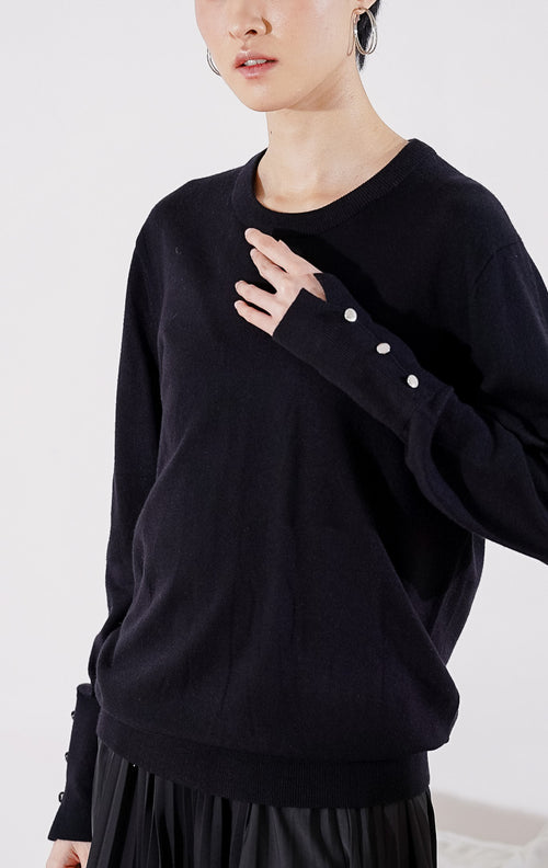 Sweater rajut wanita lengan panjang Gamora Sweater dari This Is April berwarna hitam pekat berdesain minimalis polos, kerah bulat (crewneck), dan dilengkapi aksen kancing perak elegan di bagian ujung lengan. 