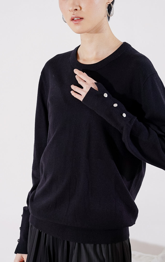 Sweater rajut wanita lengan panjang Gamora Sweater dari This Is April berwarna hitam pekat berdesain minimalis polos, kerah bulat (crewneck), dan dilengkapi aksen kancing perak elegan di bagian ujung lengan. 