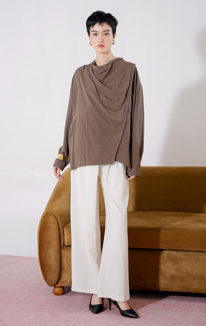 Kemeja wanita lengan panjang Georgina Top dari This Is April berwarna cokelat tua dengan desain lipatan (drape) jatuh dan detail kancing aktif menyamping asimetris di bagian depan, digantung di rak pakaian. 