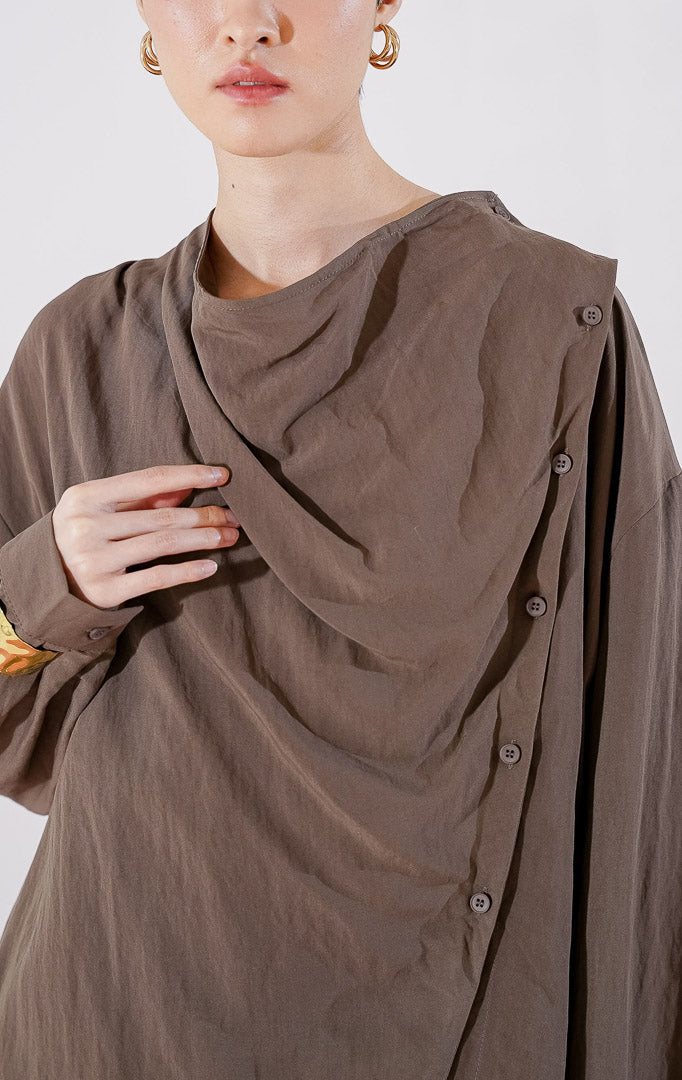 Kemeja wanita lengan panjang Georgina Top dari This Is April berwarna cokelat tua dengan desain lipatan (drape) jatuh dan detail kancing aktif menyamping asimetris di bagian depan, digantung di rak pakaian. 