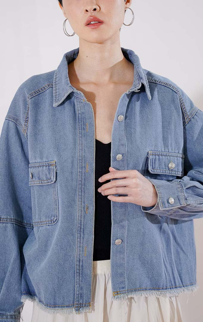 Atasan kemeja denim wanita This Is April Katanna Top warna biru dengan kerah klasik, full kancing depan, dua saku dada, dan detail pinggiran bawah raw hem 