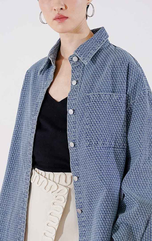Kemeja wanita lengan panjang Knox Top Denim dari This Is April berwarna biru dengan detail tekstur topstitching di seluruh bagian, dilengkapi kantong dada dan kancing depan, digantung di rak pakaian.