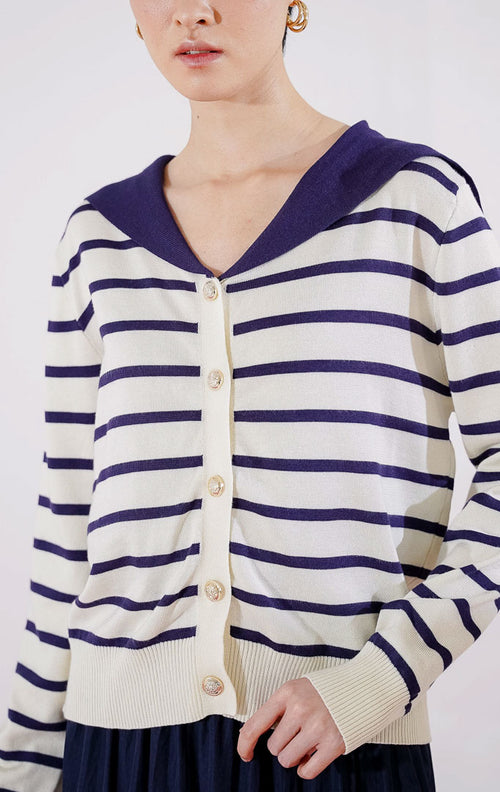Cardigan rajut wanita lengan panjang Leone Cardigan dari This Is April berwarna ivory dengan motif garis horizontal biru navy, dilengkapi kerah lebar berwarna solid navy dan kancing emas di bagian depan, digantung di rak pakaian. 