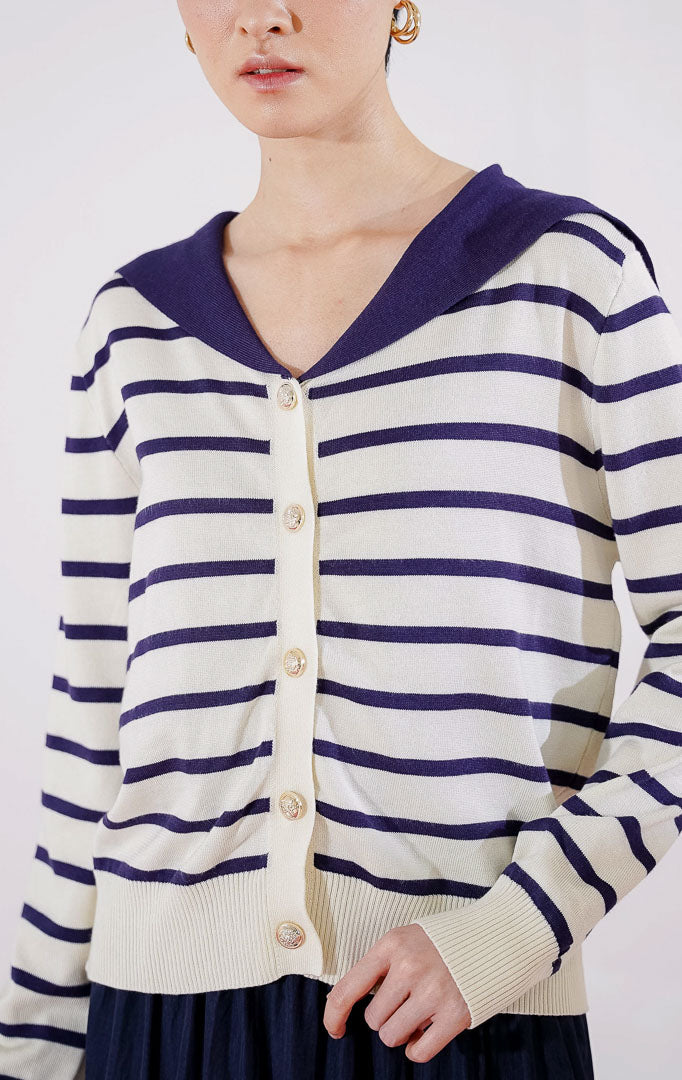 Cardigan rajut wanita lengan panjang Leone Cardigan dari This Is April berwarna ivory dengan motif garis horizontal biru navy, dilengkapi kerah lebar berwarna solid navy dan kancing emas di bagian depan, digantung di rak pakaian. 