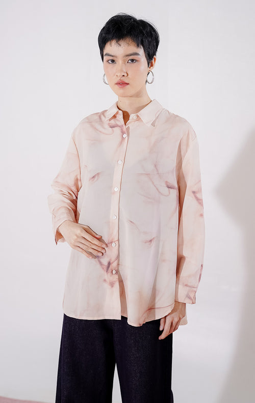 Kemeja wanita lengan panjang Manon Top dari This Is April dengan motif gradasi warna peach dan putih yang lembut, berbahan sheer menerawang dengan kancing depan, digantung di rak pakaian. 