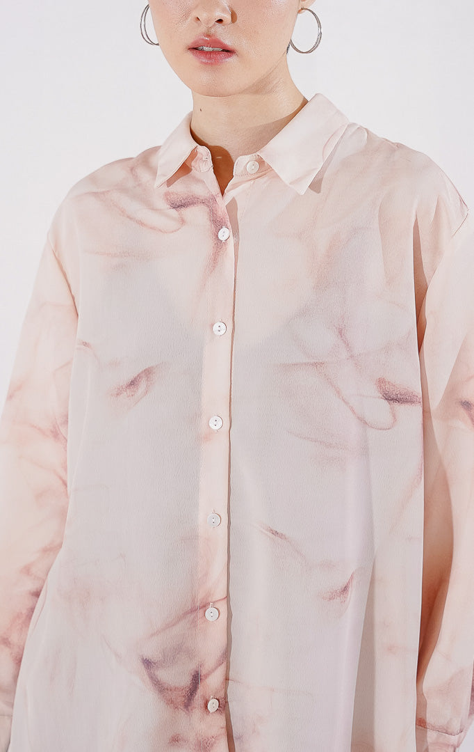 Kemeja wanita lengan panjang Manon Top dari This Is April dengan motif gradasi warna peach dan putih yang lembut, berbahan sheer menerawang dengan kancing depan, digantung di rak pakaian. 